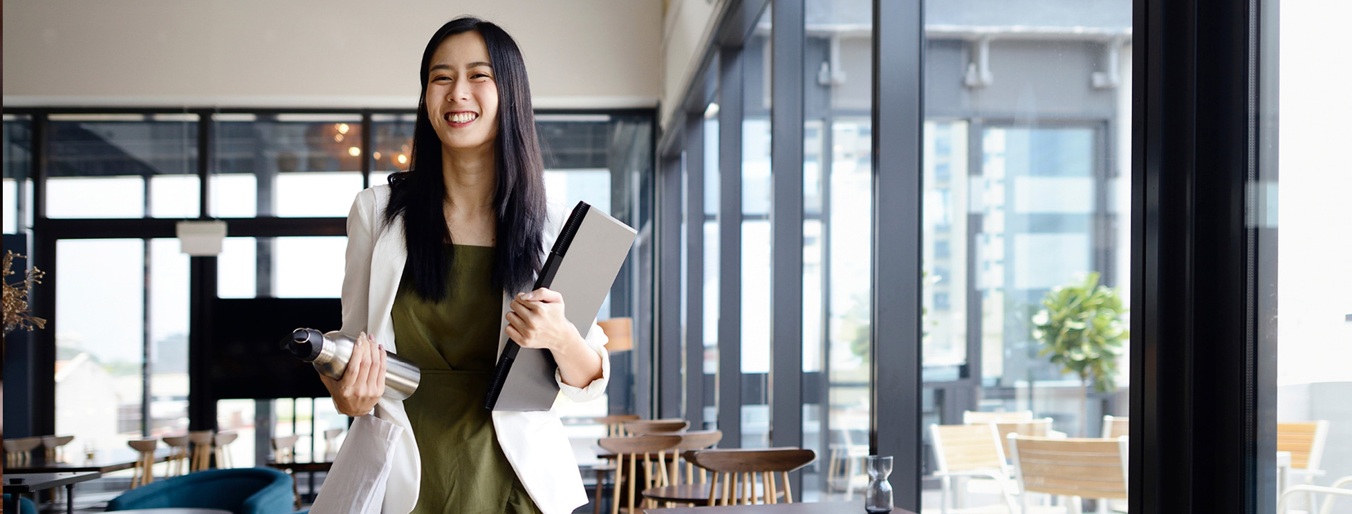 Asian woman in her twenties smiling standing in a nice office holding a laptop and water bottle.